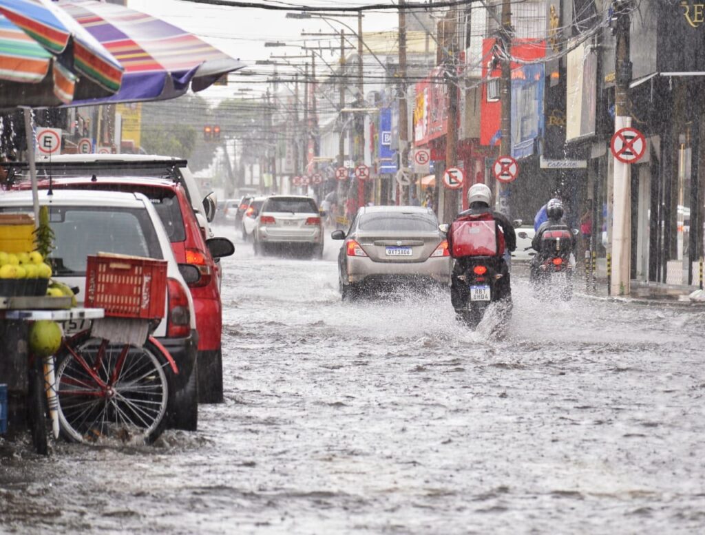 Rua alagada com carros e motos trafegando. Alerta de risco de tempestade