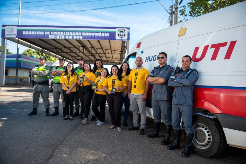 Hugol realiza ação educativa com motoristas na GO-070 - Profissionais do Hugol com camiseteas amarelas, socorristas do Samu e policiais militares, atrás deles, barreira da polícia rodoviária na GO-070 e ambulância do Hugol