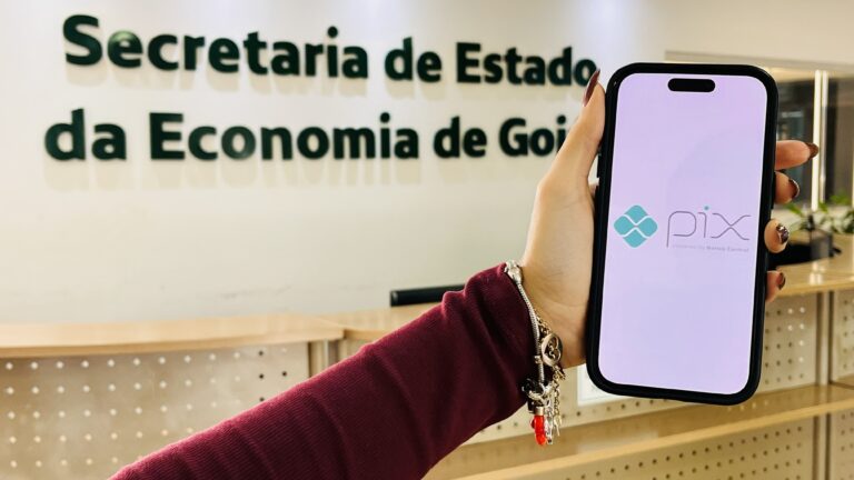 Economia vai implantar recebimento de tributos estaduais pelo Pix - Mão feminina segurando celular aberta em página do Pix, ao fundo fachada externa da Secretaria da Economia de Goiás