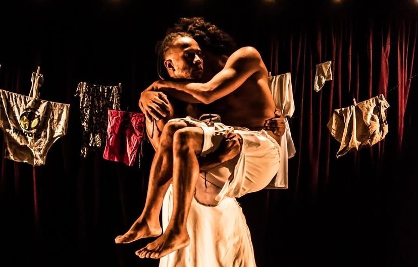 Festival Agun de Dança Negra estreia em Goiânia com imersão em arte, cultura e ancestralidade - Dois dançarinos negros no palco, o rapaz segura uma mulher em seus braços, ao fundo roupas estendidas e palco escuros