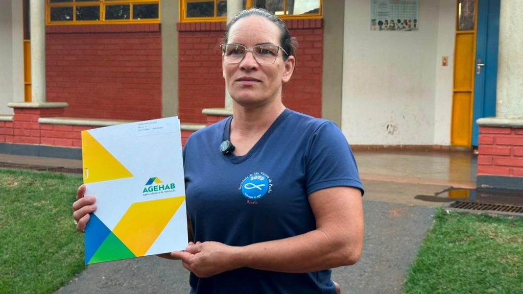 Quatro municípios goianos recebem escrituras de imóveis nesta sem - Mulher branca, de óculos, cabelos escuros presos, vestindo camiseta azul e segurando escritura